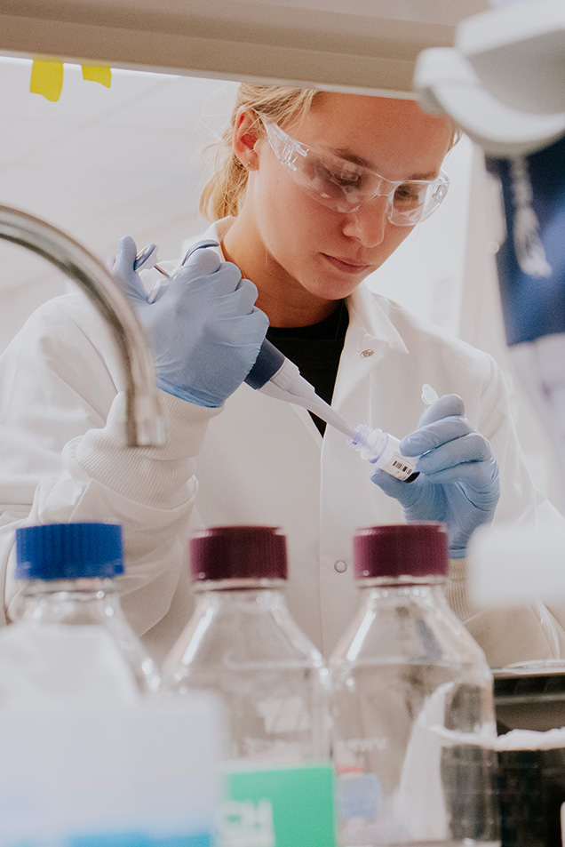 A student wearing a lab coat and gloves pours liquid into a container